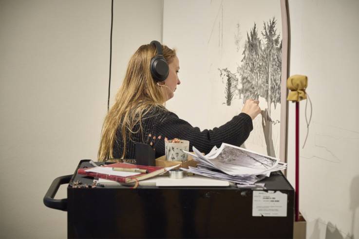 Alexandra Uppman seated at a black cart drawing a detailed forest scene on a large white wall. The cart holds papers, sketchbooks, pencils, and art supplies.  