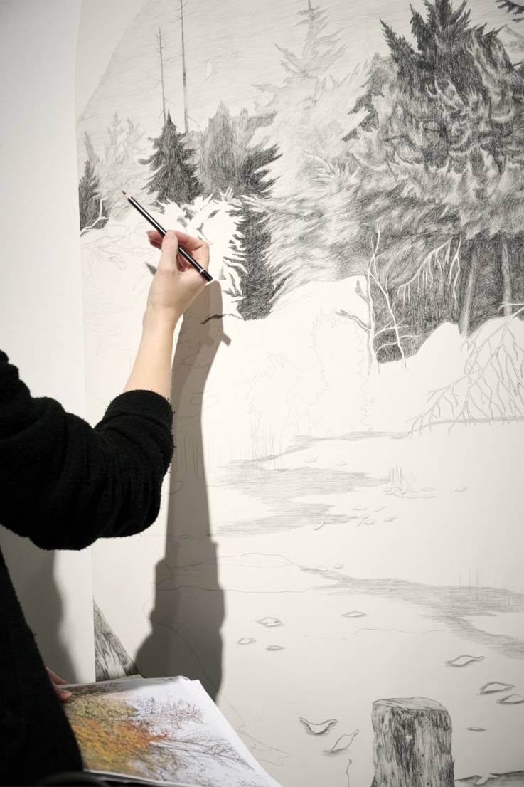 A hand holding a pencil is drawing a detailed forest landscape on a large white wall. The artwork shows trees, foliage, and a path in black and gray tones. A sketchbook with colored references rests below.