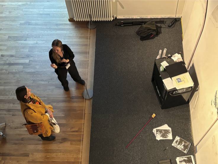 Two people standing on a wooden floor near a gray carpeted area in an art space. On the carpet is a black cabinet with papers, books, and drawing tools scattered around. A large white wall is partially visible, used for artwork.